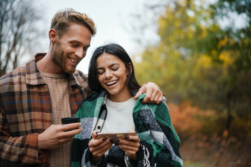 Fototapeta premium Young happy couple drinking tea and using cellphone while having picnic