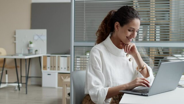 Medium shot of curly-haired Caucasian woman wearing white blouse sitting at workplace and working on laptop