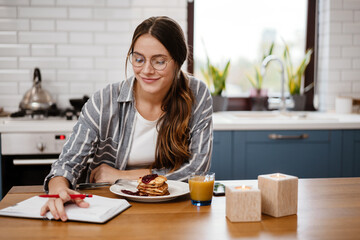 Smiling beautiful woman writing down notes while having breakfast