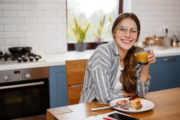 Smiling woman eating pancakes and drinking juice while having breakfast