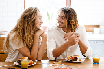 Obraz na płótnie Canvas Happy beautiful couple using cellphone while having breakfast