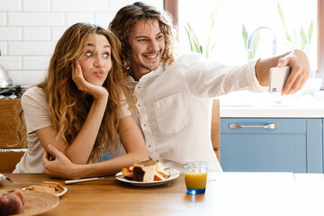 Obraz na płótnie Canvas Joyful couple taking selfie on cellphone while having breakfast