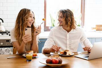 Cheerful couple using cellphone and laptop while having breakfast