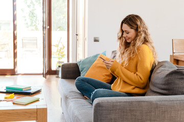 Happy beautiful girl smiling and using cellphone while resting on couch