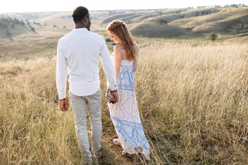 Multiracial couple in love walking on the summer meadow holding hands, back view