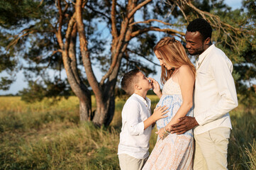 Happy diverse family spends time outdoors together. Pregnant wife, care husband and school age boy on the meadow in warm summer day