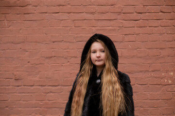 Naklejka premium portrait of cute brunette teen girl in black fur coat on the background of brown brick wall . Cosiness, fashion, style