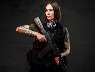 Stylish and martial woman with short haircut and tattooed arm poses holding ak74 rifle in dark background.