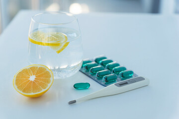 Glass with lemon water, blue capsules pills and termometer lie on white table in hospital. Vitamin drink and medicine for recovery from virus illness. Healthcare concept.