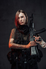 Fototapeta na wymiar Dreseed in dark armour military woman with short haircut holds rifle with tattooed and bandaged hands in dark background.