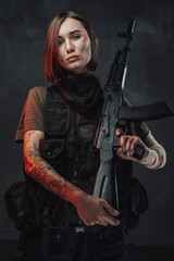 Fototapeta premium Seductive female soldier with tattoo and bandaged hand poses in dark foggy background holding rifle on her shoulder.