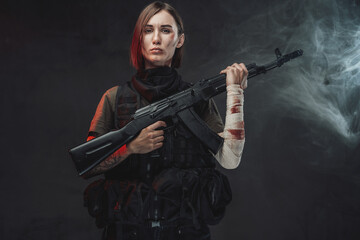 Dressed in dark armour military woman with short haircut and bandaged hand holds her assault rifle in dark smokey background.