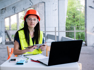 Woman architect in protective helmet and goggles working on personal computer on new project at construction site
