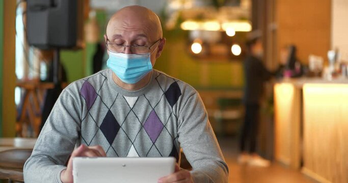 Senior Man In Medical Protective Mask And Glasses Sits At The Table In Restaurant And Uses Tablet Pc. Covid Concept Health And Safety, N1H1 Coronavirus Quarantine, Next Wave Covid Virus Protection