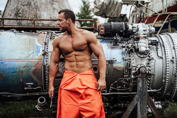 Fototapeta na wymiar Portrait of a mechanic macho in orange clothing with muscular build looking away and posing near engine of a airplane.