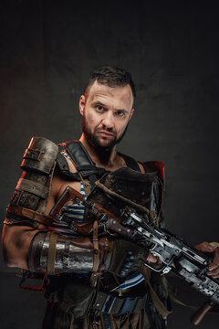 Portrait of a post apocalyptic bearded guy dressed in ragged armour holding custom shotgun in dark background.