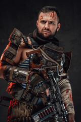 Brutal and bearded apocalyptic fighter in dirty and ragged armour poses in dark background holding his custom shotgun.