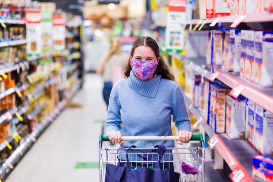 Young female shopper walking down grocery store aisle wearing home made face mask looking at camera