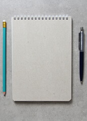 A spring notebook with a sheet of craft paper A5 with a ballpoint pen and simple green graphite pencil on light grey concrete background. Empty space for text.