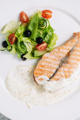 Piece of baked salmon with sour cream sauce with green salad on a white plate top close-up view
