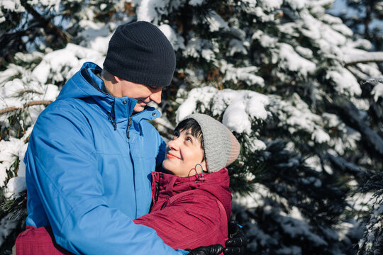 Middle Age Couple Hug At The Snowy Winter Park. A Man And A Woman In Blue And Red Jackets Having Fun Among Fir Trees Covered With Snow. Love, Togetherness And Holidays Concept.