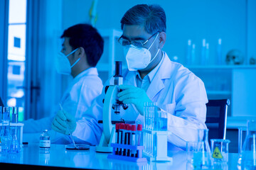 Fototapeta premium Asian medical doctor scientists in lab gown and face protective mask working with microscope and test tubes in clinical laboratory. virus pandemic concept. healthcare and medicine concept. 