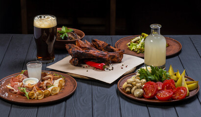 Food set with grilled pork ribs, glass of dark beer, pickled vegetables, bacon, salad, and moonshine  on black wooden background