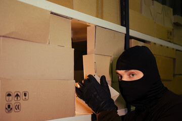 A thief in a black mask steals a box of goods in a warehouse in the dark. Concept of problems with...