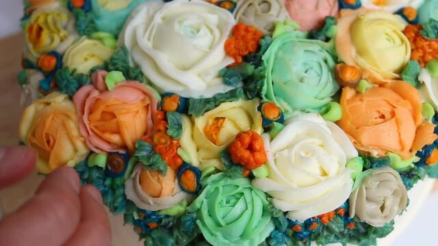 The pastry chef touches the cream rose petals with his bare finger. Bright elegant cake decorated with flowers of roses, Ranunculus and Rubus chamaemorus. Top view, close-up, contains people, real tim