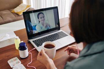 Fototapeta na wymiar Young woman drinking cup of hot tea when sitting at table with laptop and video calling her doctor