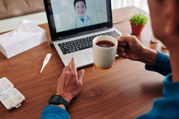 Fototapeta na wymiar Sick man drinking cup of warm black tea when talking to his doctor via telemedicine platform