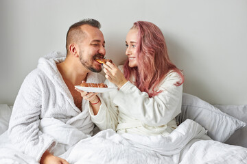 Obraz na płótnie Canvas female treats husband with croissants eclairs, caucasian couple on bed in the morning, love concept