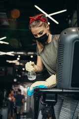 Young female worker disinfecting cleaning and weeping expensive fitness gym equipment with alcohol sprayer and cloth. Coronavirus global world pandemic and health protection safety measures.