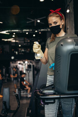 Young female worker disinfecting cleaning and weeping expensive fitness gym equipment with alcohol sprayer and cloth. Coronavirus global world pandemic and health protection safety measures.