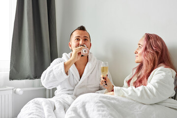 Obraz na płótnie Canvas beautiful couple have romantic time on bed drinking champagne, caucasian man and woman in bathrobes drink beverage, have talk, celebrating honeymoon