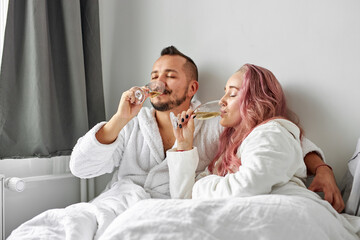 Obraz na płótnie Canvas beautiful couple have romantic time on bed drinking champagne, caucasian man and woman in bathrobes drink beverage, have talk, celebrating honeymoon
