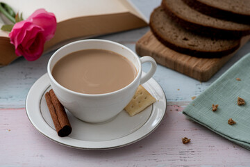 Sliced whole wheat bread, coffee and cream, and an open book marked with pink rose, on aged wooden table, decorated with cinnamon stick, a cracker, and a pink rose- afternoon coffee at home concept