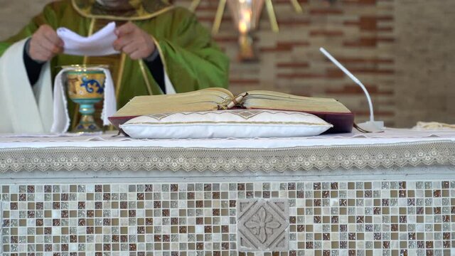 Close-up view of catholic priest standing next to church altar, reading open Holy Scripture and preparing Holy Eucharist for ceremony. Pastor in green mantle using chalice, sacramental bread and wine