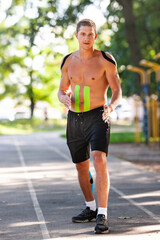 Full length portrait of handsome young caucasian professional bodybuilder with elastic tapes on body running at stadium. Man with muscular naked torso looking at camera. Workout concept.