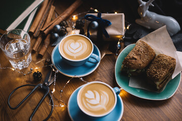 Two delicious cups ofbcappuccino coffee standing on the rustic wooden table among Christmas decorations, fireflies, wrapped gift, scissors, cinnamon sticks, deer toy, with sandwiches on the background