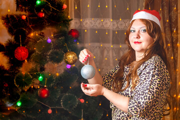 Naklejka premium A red-haired woman in a Santa hat by a Christmas tree with a decoration ball in her hands.