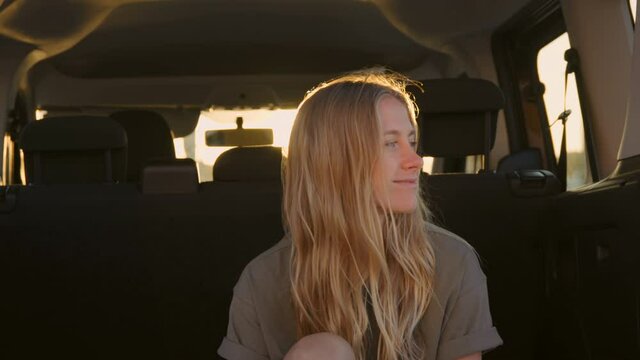 Portrait of beautiful authentic young woman look at camera, smile and laugh, relaxed and real emotions and feelings. Woman sit in back of car at sunset. Road trip concept