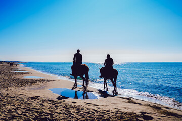 silhouette of two people riding two horses on the beach on a relaxing winter afternoon