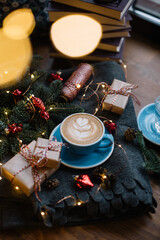 Delicious fresh festive morning cappuccino coffee in a ceramic blue cup on the warm cover with little wrapped gifts, red ornamentals, fireflies and spruce branches