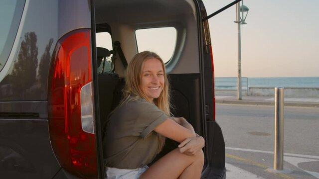 Young surfer woman sit in back of car and enjoy view of sunset on empty beach. Summer day, small weekend trip to seaside, staycation concept