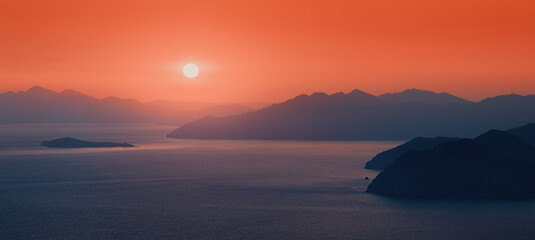 Moody sunset, the distant sun sets behind the silhouettes of blue mountains on the horizon, washed by the waters of the Mediterranean sea