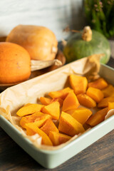 Baked pumpkin sliced in small pieces  in a baking dish on a wooden background with different  fresh pumpkins. Selective focus, copy space. Vegan healthy food.