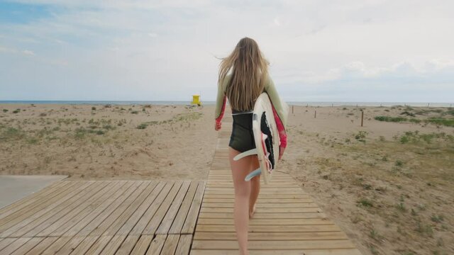 Cinematic shot of amazing beautiful athletic female surfer walk on wooden boardwalk towards water waves on empty holiday beach. Surfing woman, professional sports equal representation. Inspiring