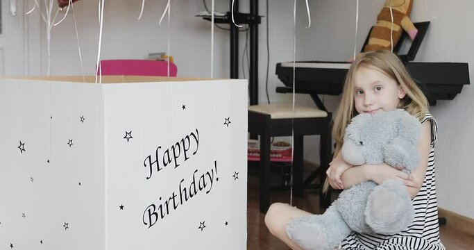 Cute baby girl hugs a plush taddy bear toy. A large cardboard box with birthday gifts stands on the floor.