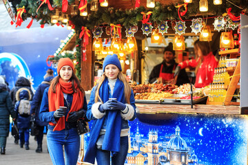 Fototapeta na wymiar two young girls in red and blue hats and scarves walk around the Christmas market, drink coffee, talk and smile